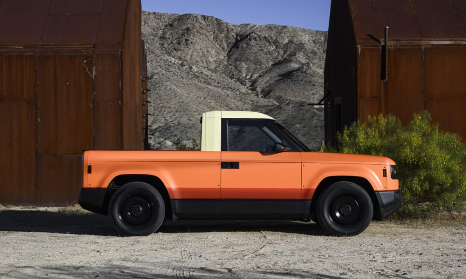 Orange Slate electric pickup truck parked outdoors between industrial structures, representing the company’s upcoming customizable EV supported by new funding.