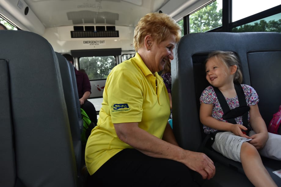 A school bus driver from Student Transportation of America smiles and talks with a young girl buckled safely into her seat on the bus.