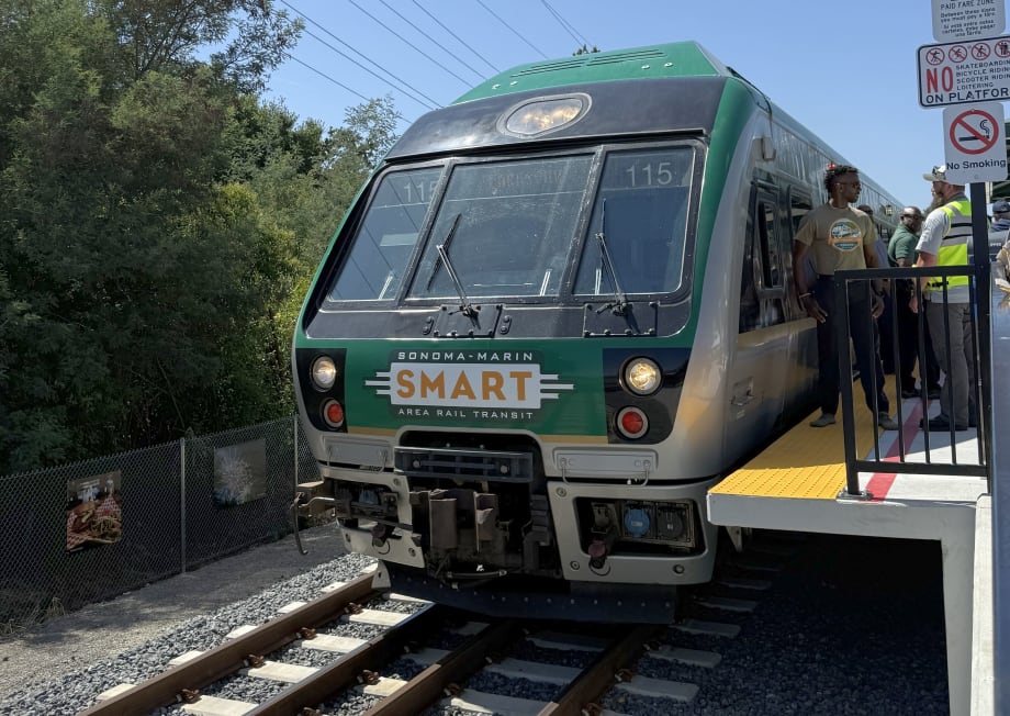 A SMART (Sonoma-Marin Area Rail Transit) train stopped at a platform during a station opening event, with people standing beside the train and signage posted near the edge of the platform.