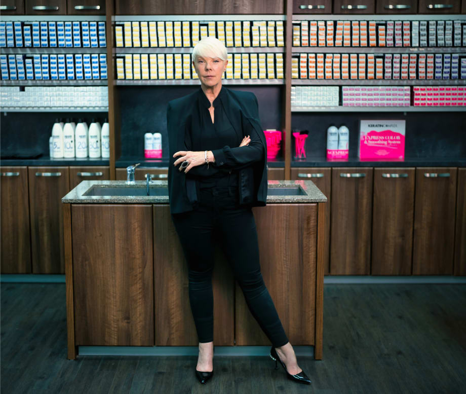A woman in black and with short blonde hair, with arms crossed, standing in front of shelves of beauty products.