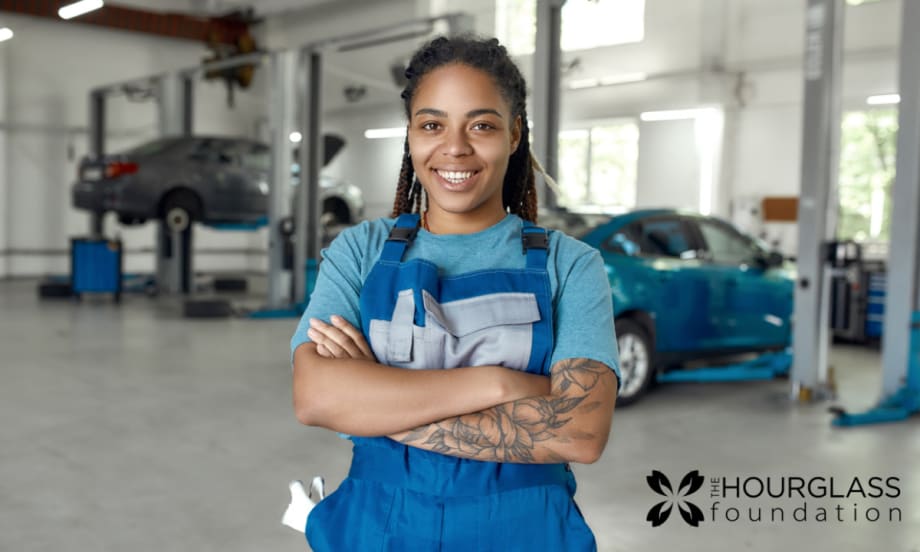 A woman in a blue shirt and apron stands in an automotive suite with her arms crossed.