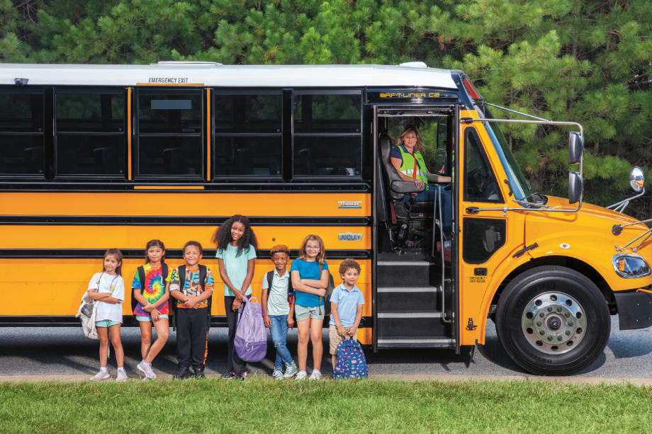 A group of students stands in front of a Thomas Built Jouley electric school bus. The door is open with the bus driver smiling for the camera.
