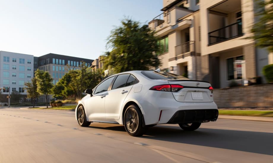 A 2026 Toyota Corolla drives on the road towards a glass building.