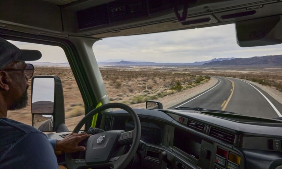 Truck driver navigating a winding desert highway, viewed from inside the cab of a Volvo semi-truck, with mountains and open landscape visible through the windshield.