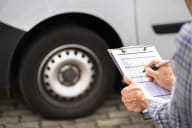 A person holding a clipboard and writing on an inspection checklist beside the wheel of a large white vehicle, likely conducting a fleet or safety inspection.