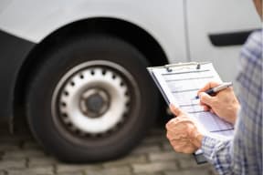A person holding a clipboard and writing on an inspection checklist beside the wheel of a large white vehicle, likely conducting a fleet or safety inspection.