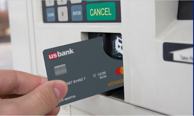 A person inserts a U.S. Bank Voyager Fleet card into a gas pump card reader. The gray credit card features the U.S. Bank logo, chip, and Mastercard logo, while the fuel pump display shows buttons for “Enter” and “Cancel.”