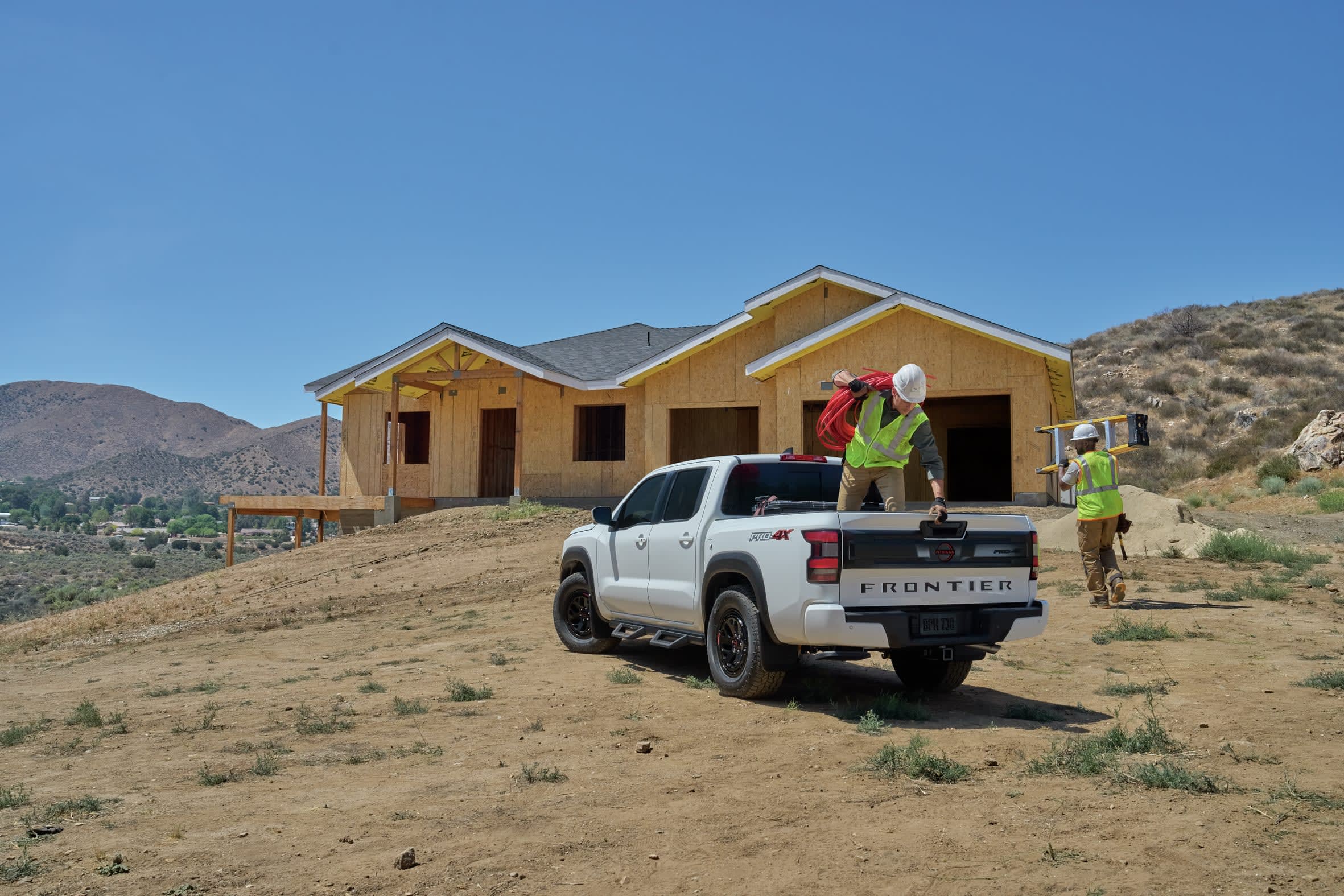 White Nissan Frontier pickup truck parked on a construction site with a partially built house in the background. Two workers in hard hats and safety vests are near the truck and the house, one carrying coiled red wire from the truck bed and another carrying a ladder.