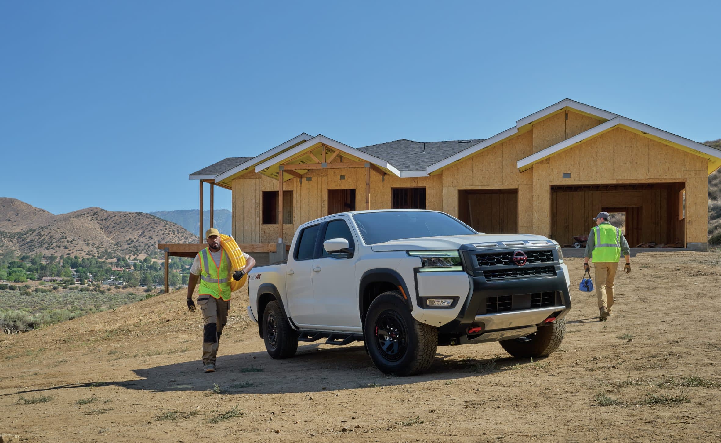 A white Nissan Frontier pickup truck is parked on a dirt lot in front of a house under construction, with two construction workers in high-visibility vests walking nearby on a sunny day.