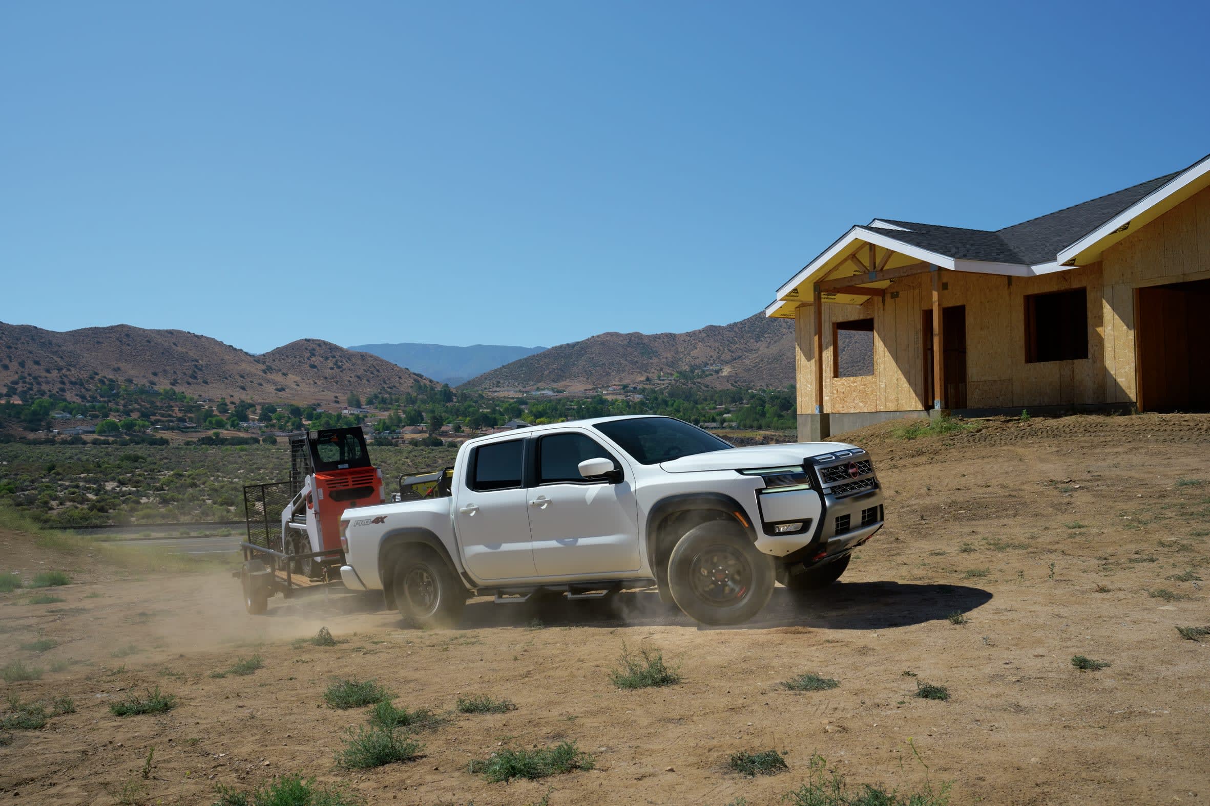 A white Nissan Frontier pickup truck tows a trailer with an orange and black compact construction vehicle across a dusty construction site, with a house under construction in the background and rolling hills under a clear blue sky.