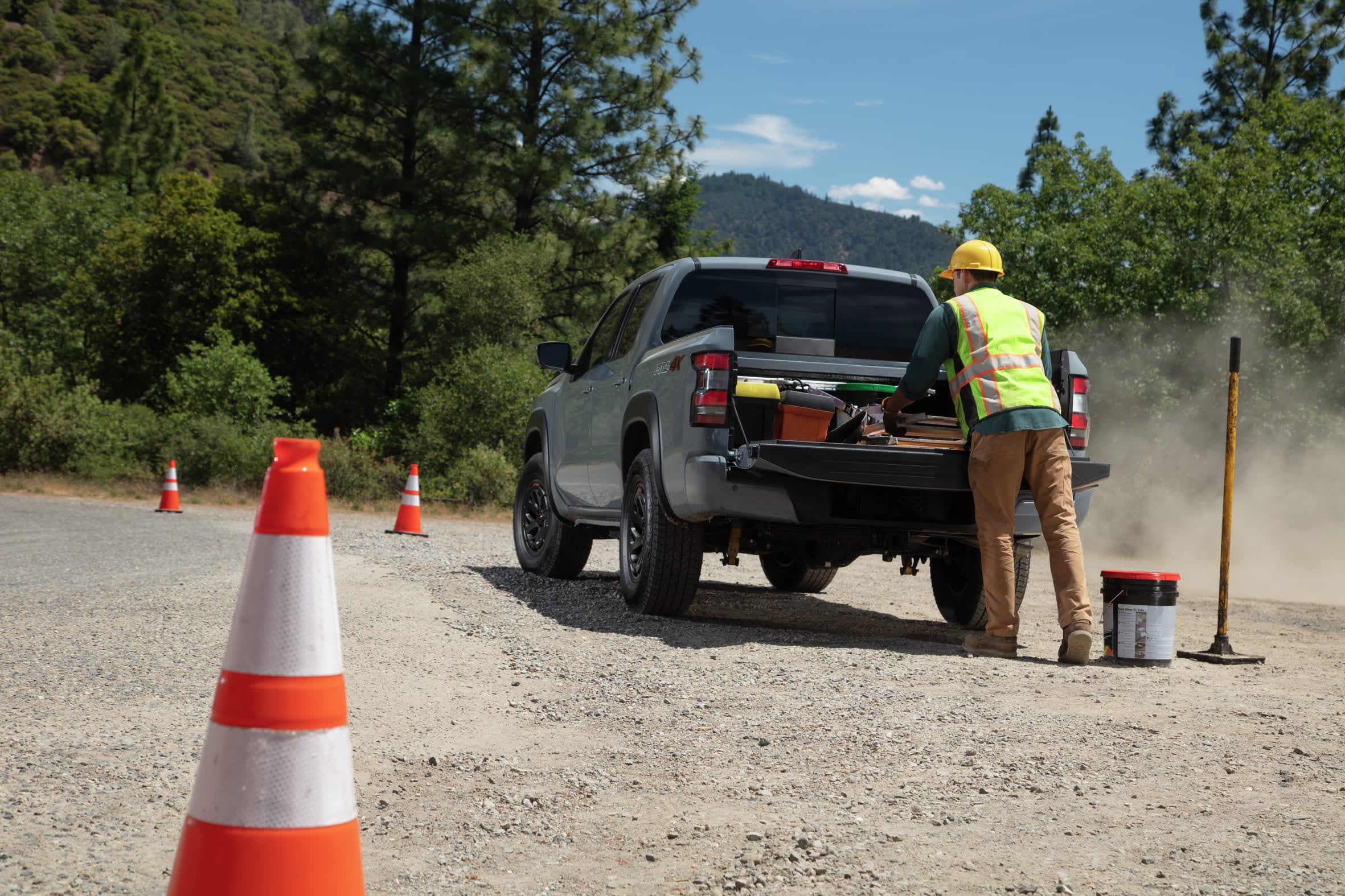 A person in a reflective vest and hard hat loads tools into the open tailgate of a grey Nissan pickup truck on a dusty, gravel road.