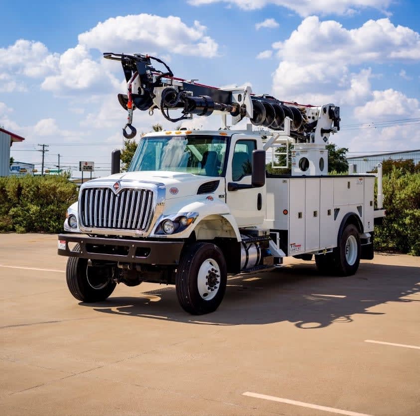 White utility truck with an extended boom crane mounted on top, parked outdoors in a paved lot with buildings and green bushes in the background under a bright blue sky with fluffy white clouds.