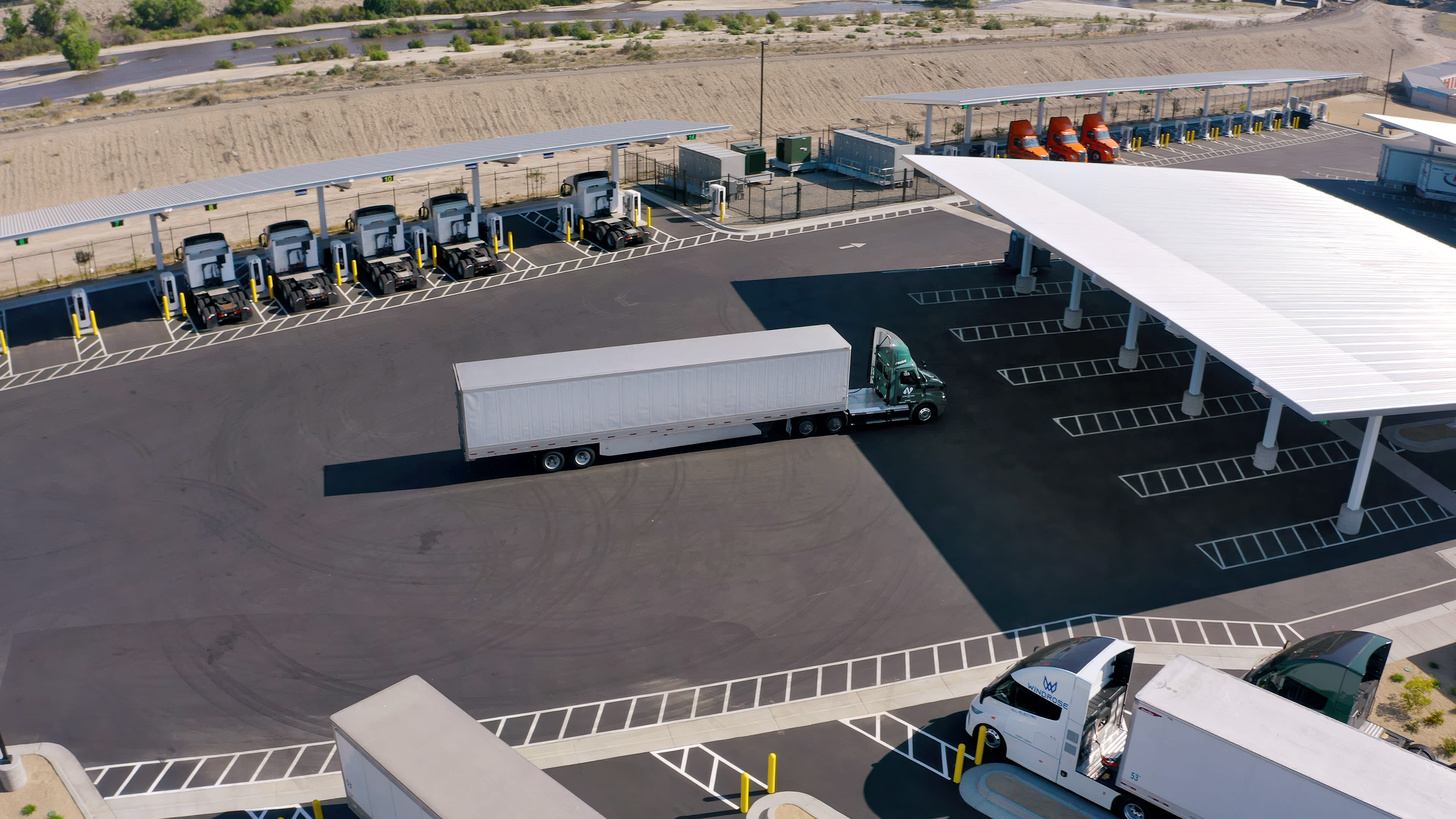 Aerial view of a Greenlane EV charging center: semi-trucks lined at loading docks, trailers in a paved freight yard beside a large covered parking canopy.