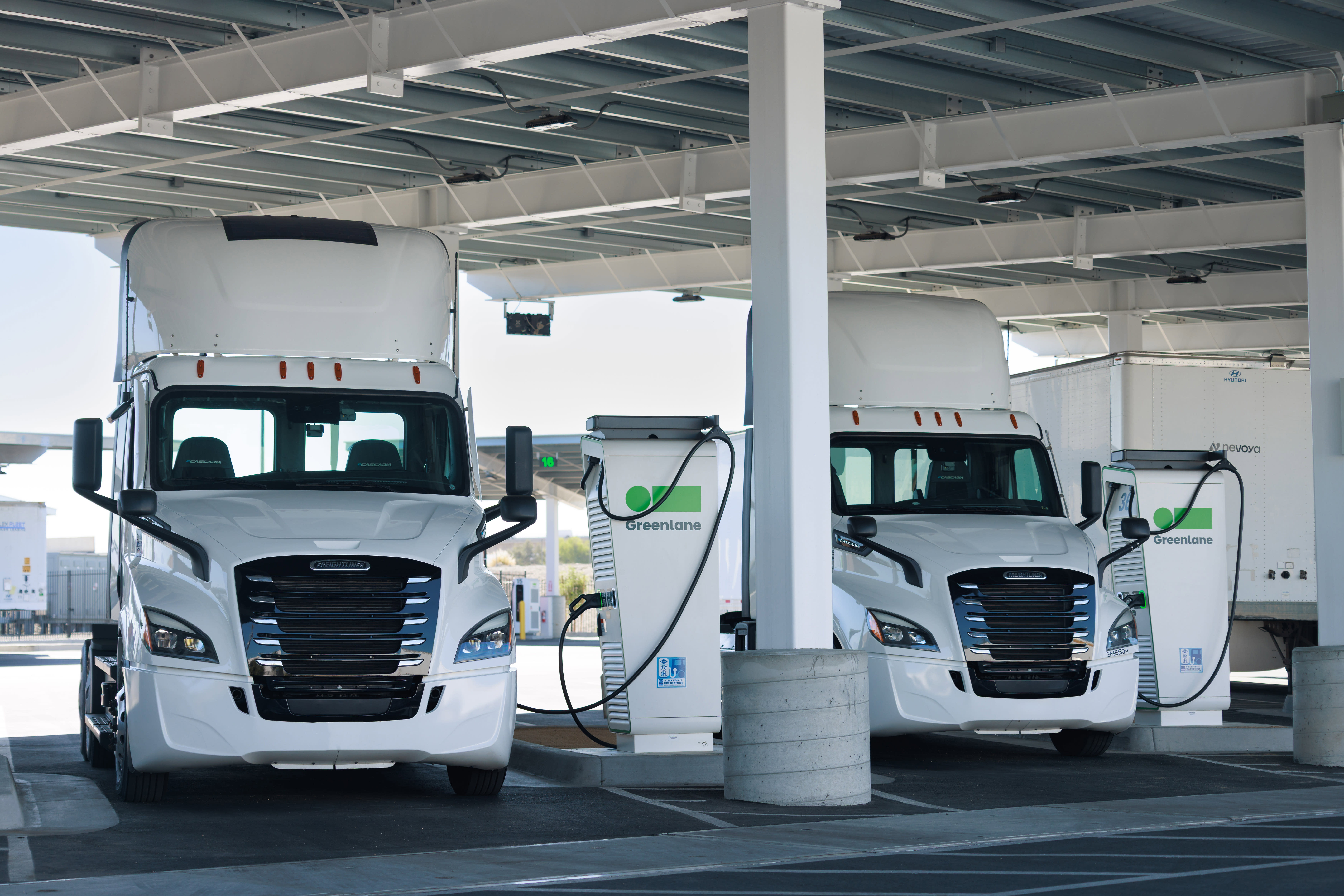 Two white electric semi-trucks at a Greelane charging station under a covered depot, showcasing sustainable fleet technology and EV freight infrastructure with charging stations.