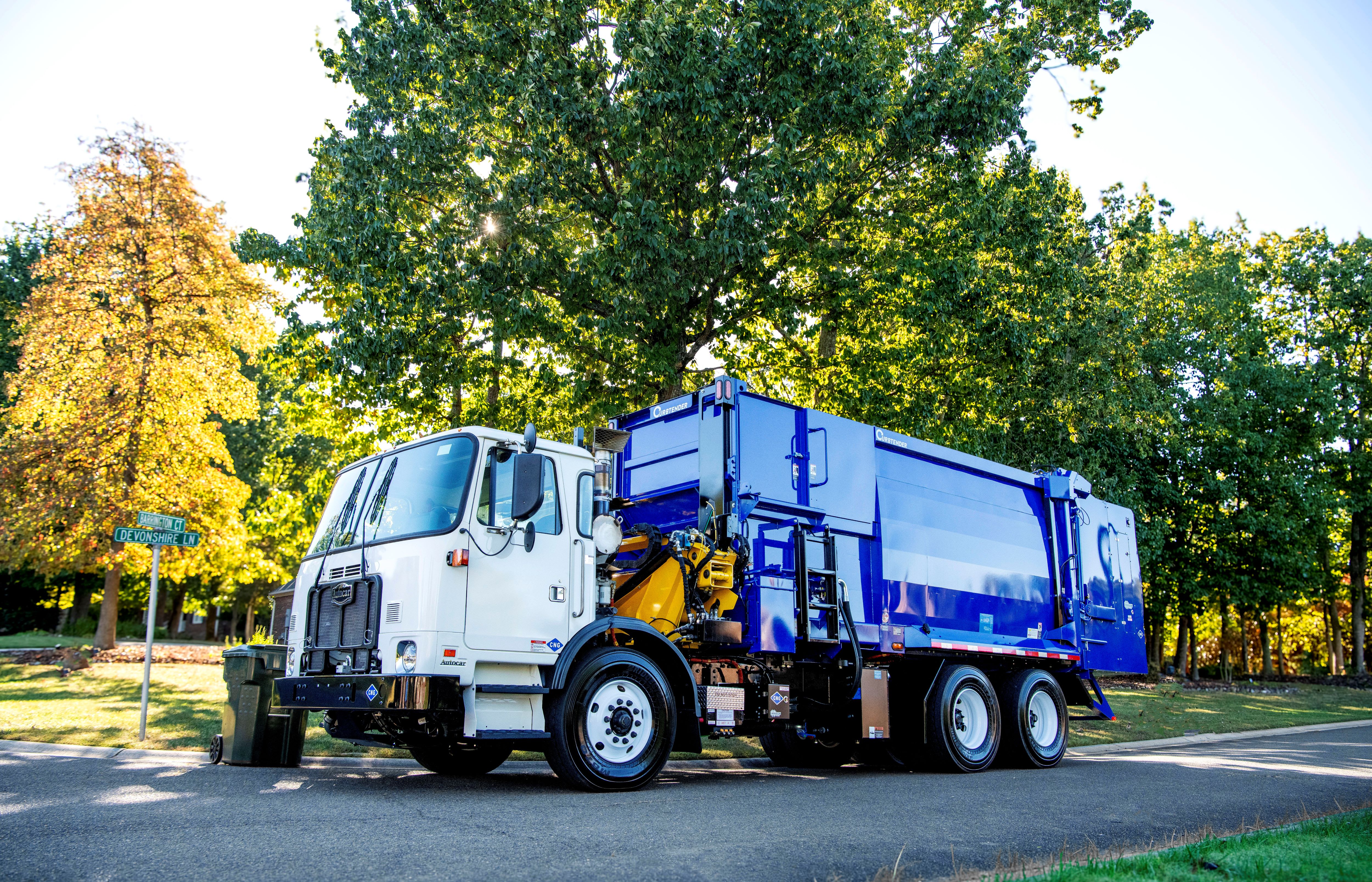 Blue Hegaxon Agility refuse truck parked on a sunlit, tree-lined street, highlighting waste collection services and urban recycling in a green neighborhood.