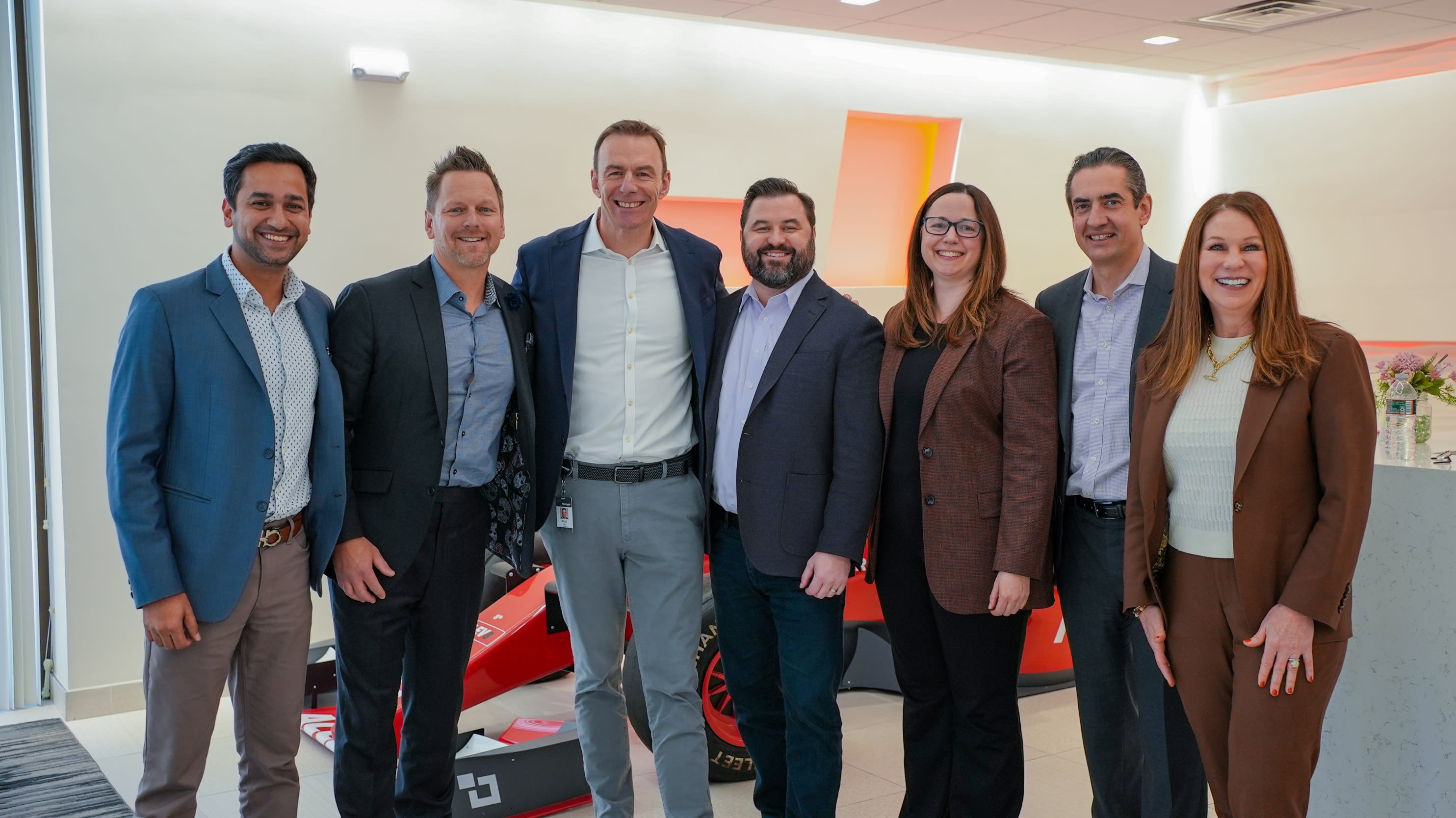 Seven business professionals in smart attire pose smiling beside a red sports car in a bright modern showroom. Merchants Fleet team photo showcasing automotive partnership.