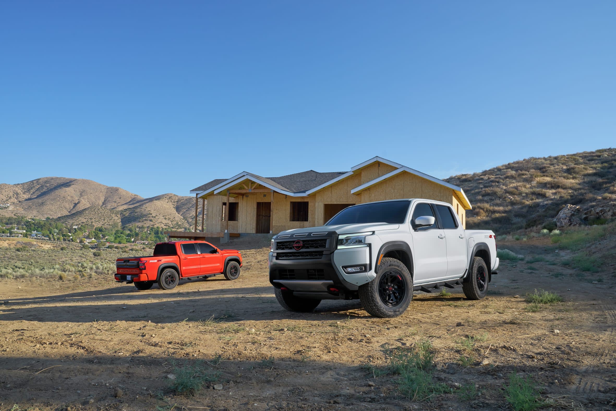 Two Nissan pickup trucks, one white and one red, are parked in a dusty, semi-arid landscape in front of a house under construction.