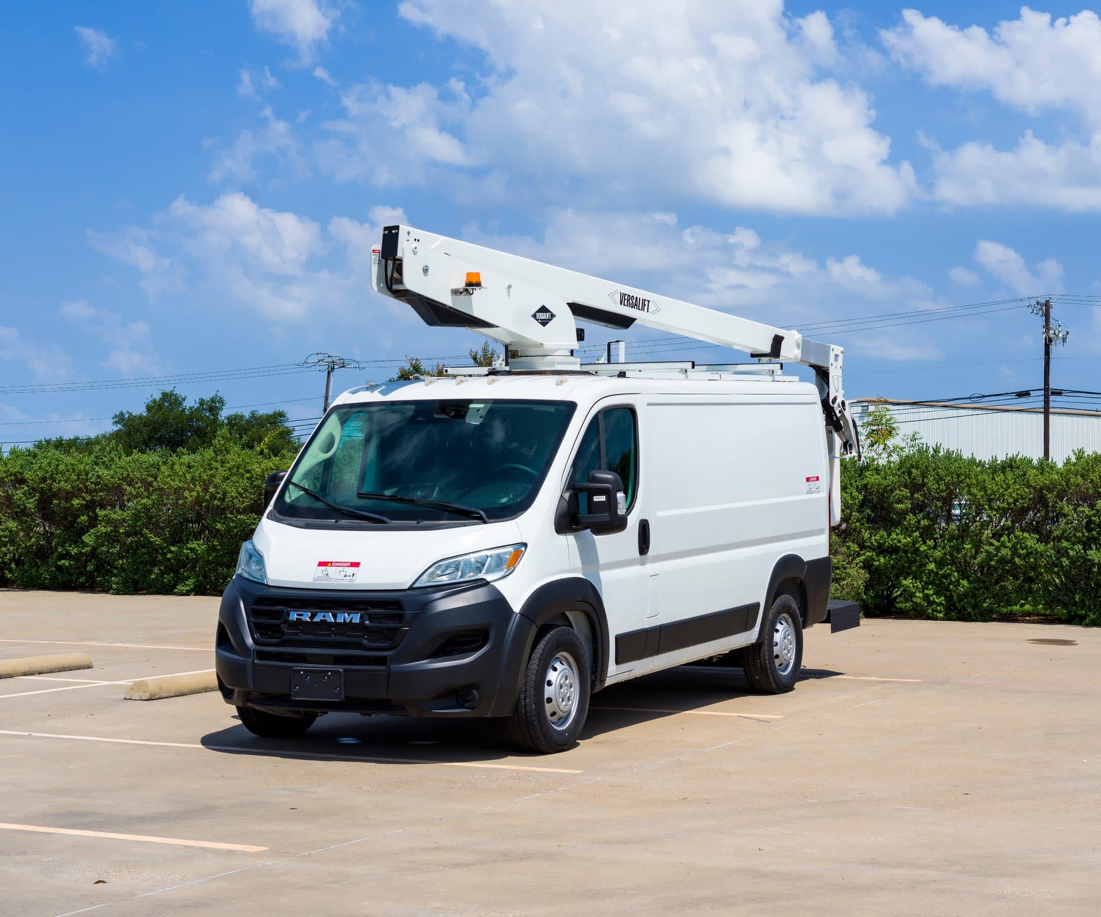 White Ram ProMaster van with a utility boom lift mounted on the roof, parked in an empty outdoor parking lot under a blue sky with white clouds.