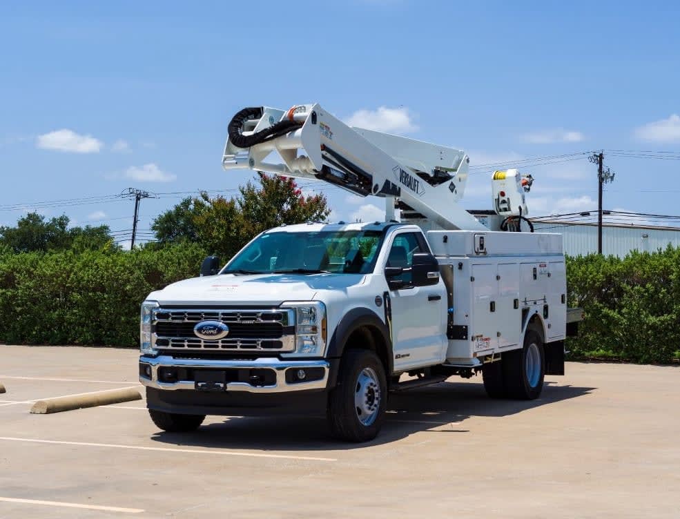 White Ford utility truck with an extended bucket lift arm parked in an empty asphalt lot with green bushes and a building in the background under a bright blue sky with scattered clouds.