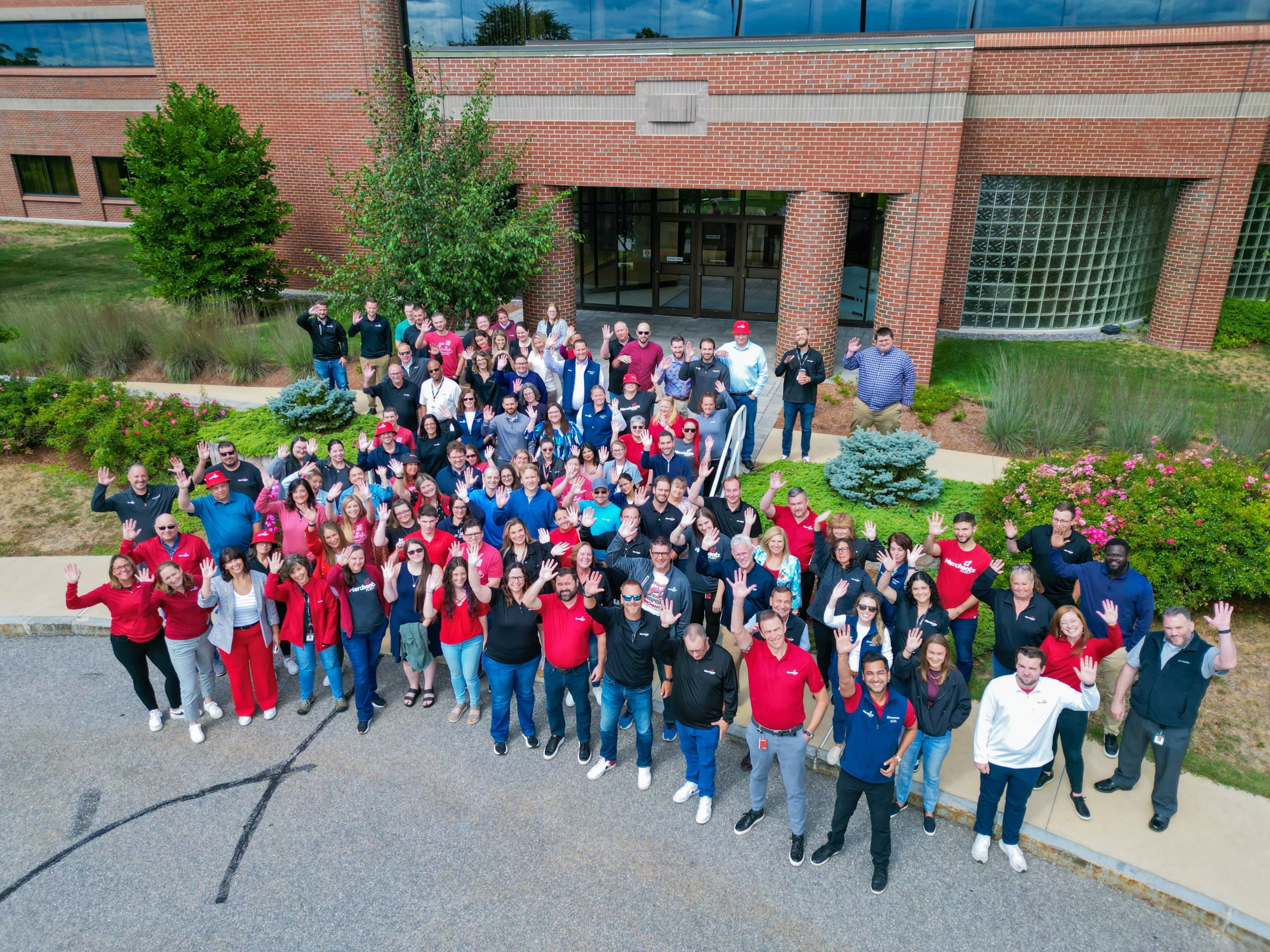 Merchants Fleet team photo. Large diverse group of employees gathered outside a modern brick office building, smiling and waving for a spirited company team photo.