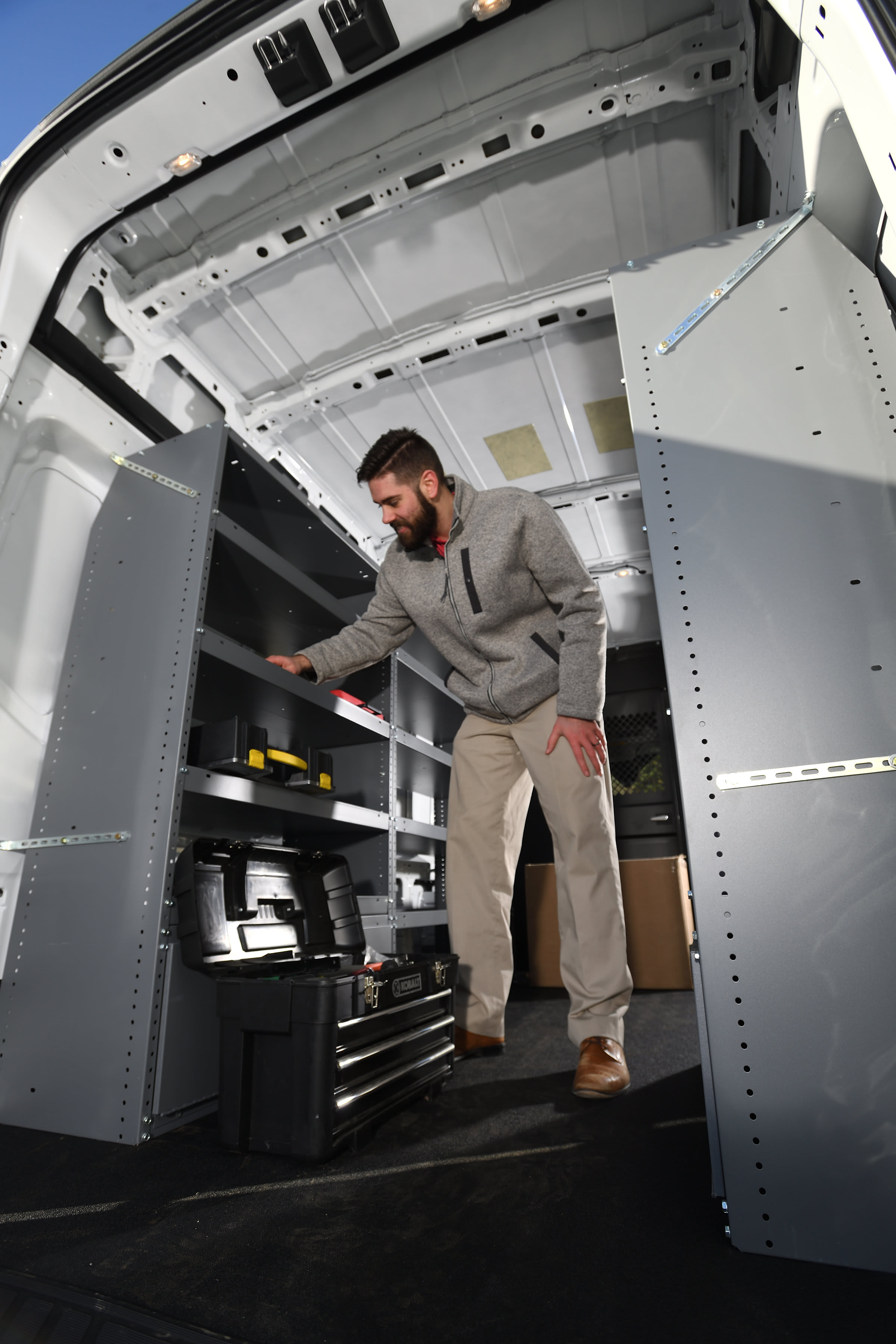 Technician in work jacket organizing tools inside a white service van equipped with modular shelving and drawers—mobile workshop ready for on-site repairs and service.